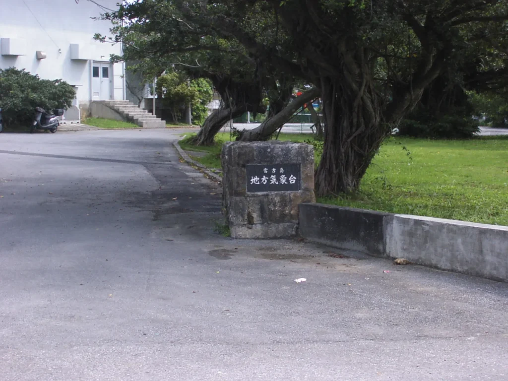 「宮古島 地方気象台」と刻まれた石造りの門柱。敷地内には南国らしい立派な樹木(ガジュマルなど)が枝を広げている。